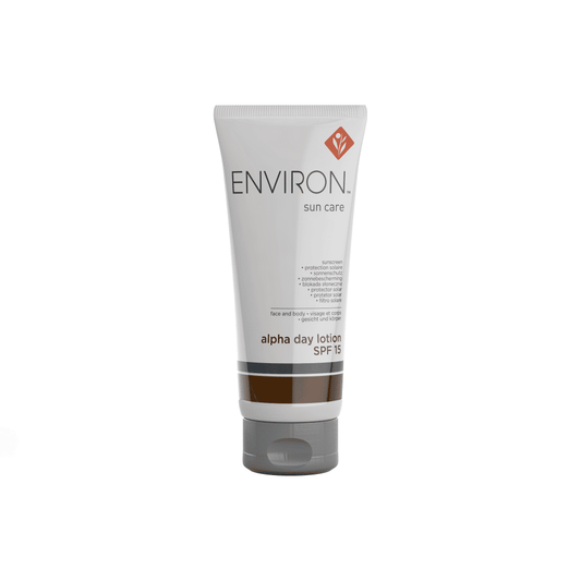 A white tube of Environ Alpha Day Lotion SPF 15 labeled "SPF 15." The tube features a brown stripe near the bottom and a logo with a red diamond shape near the top. This sunscreen claims it offers sun protection and promotes skin health.